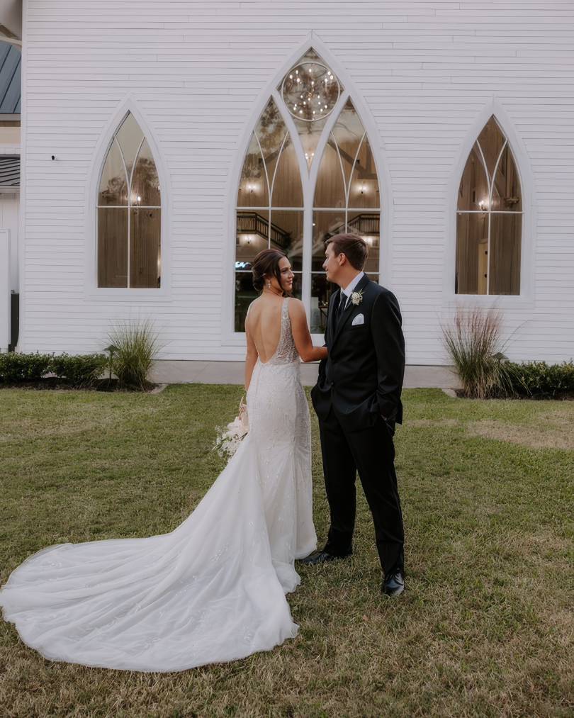 bride and groom looking at each other in bride's justin alexander wedding dress in front of the white farmhouse-style building at the springs wallisville